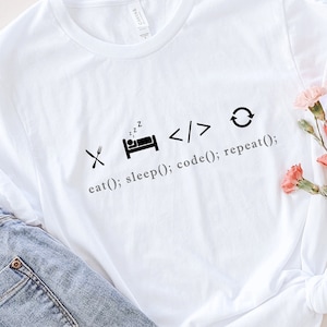 May include: White t-shirt with the text "eat(); sleep(); code(); repeat();" in black. The shirt is paired with light blue jeans and a straw hat. Pink flowers are arranged on the side.