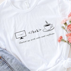 Puede incluir: Camiseta blanca con el texto "Fueled up with code and caffeine" y gráficos de un monitor de computadora, corchetes de código y una taza de café. La camiseta se combina con vaqueros de lavado claro y un sombrero de paja. Flores rosas dispuestas a un lado.