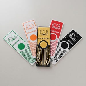 Può includere: Una collezione di cinque segnalibri rettangolari con l'icona di un libro e la parola "BOOKMARK". I segnalibri sono verdi, arancioni, rossi, neri e dorati. Il segnalibro dorato presenta un design a circuito stampato.