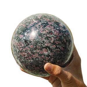 May include: A polished, spherical gemstone with a speckled pattern of dark gray, black, and deep red. The stone is held in a hand, showcasing its smooth, rounded form. The overall effect is of a natural, decorative object.