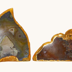 Può includere: Due fette di agata lucidata con venature naturali in tonalità grigie, marroni e bianche. Ogni pietra presenta una forma irregolare e frastagliata con un bordo metallico spesso e testurizzato di colore dorato applicato sul perimetro esterno.