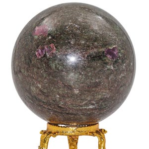 Peut inclure: Une sphère de pierre polie, de couleur verte et marron mouchetée, ornée de grappes de cristaux roses. La sphère repose sur un support décoratif en métal doré orné de trois pieds. L'ensemble suggère un objet décoratif.
