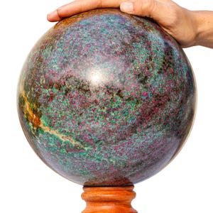 Può includere: Una grande sfera lucidata con una superficie screziata di tonalità verdi, bordeaux e marroni. La sfera poggia su un piccolo supporto di legno. Una mano poggia delicatamente sulla parte superiore della sfera. La sfera sembra essere di pietra.