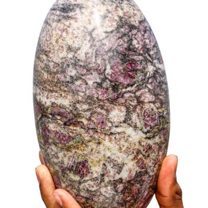 May include: A large, oval-shaped, pink and grey gemstone with a swirling pattern. The stone is being held in a person's hand.