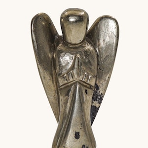 Peut inclure: Une figurine d'ange en métal argenté avec une surface lisse et réfléchissante et une tête stylisée sans visage. La silhouette présente de grandes ailes déployées et les mains jointes en prière, avec des taches sombres et usées visibles.