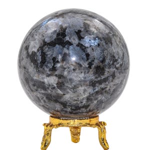 May include: A spherical gemstone with a mottled grey and black pattern, displayed on a small, ornate gold-coloured metal stand. The gemstone is a decorative item. The stand has three legs. The sphere is a decorative item.