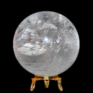 Puede incluir: Una esfera de cristal de cuarzo transparente, apoyada sobre un soporte de metal dorado con tres patas ornamentadas. La esfera tiene fracturas e inclusiones internas, sobre un fondo negro.
