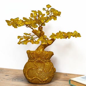 May include: A decorative tree sculpture featuring translucent yellow crystal leaves attached to twisted gold-coloured wire branches. The tree is anchored in a textured, gold-coloured base shaped like a drawstring money bag with a circular emblem.