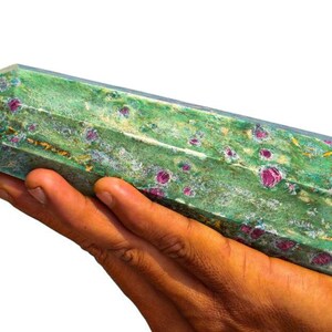 May include: A green and red ruby crystal wand. The crystal is held in a hand. The rectangular crystal has angled edges. The green stone is speckled with red ruby inclusions.