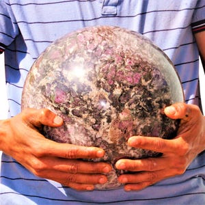 May include: A large, polished sphere of pink and grey stone with a marbled pattern. The sphere is being held in the hands of a person.
