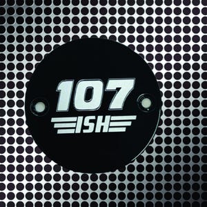 May include: A black circular emblem with the number "107" in white, and the word "ISHE" below it, against a silver perforated metal background. The emblem has two small silver rivets. The background has a pattern of evenly spaced black dots.