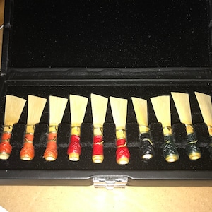May include: A black case with a velvet lining holds a set of 10 oboe reeds. The reeds are arranged in two rows of five, with the top row featuring red and orange colored reeds and the bottom row featuring black and green reeds.