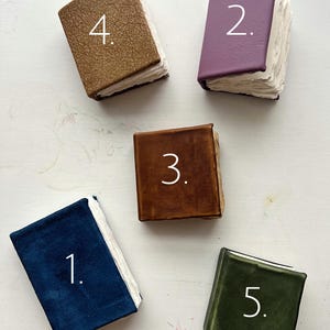 May include: An overhead shot of five small, handmade books with textured covers in various colors: navy blue, purple, brown, tan, and olive green. Each book has a white page block and a number in white on the cover.