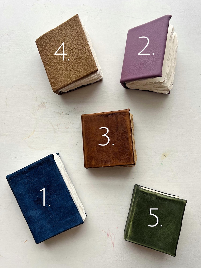 May include: An overhead shot of five small, handmade books with textured covers in various colors: navy blue, purple, brown, tan, and olive green. Each book has a white page block and a number in white on the cover.