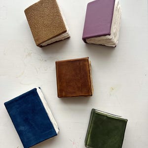 May include: An overhead shot of five miniature handmade books. The books have covers in various colors: brown, purple, brown, blue, and green. The pages are white and appear to be deckle-edged. The books are arranged on a white surface.