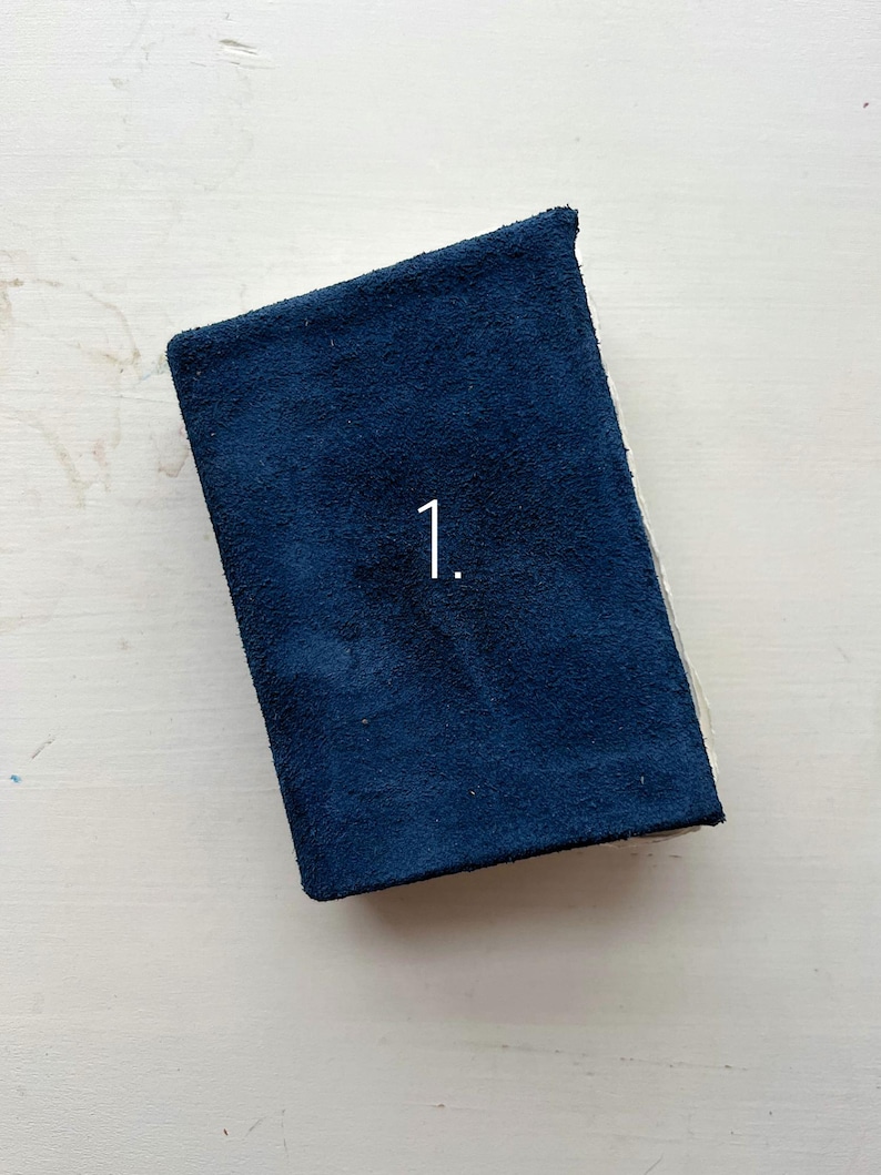 May include: A navy blue, rectangular book or journal with the number "1" printed on the cover. The cover has a textured, suede-like appearance. The book is resting on a white surface.