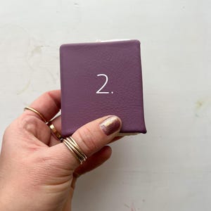 May include: A small, rectangular, purple box with the number "2" in white. The box is held in a hand with multiple gold rings on the fingers. The background is a white surface.
