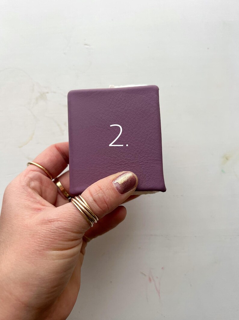 May include: A small, rectangular, purple box with the number "2" in white. The box is held in a hand with multiple gold rings on the fingers. The background is a white surface.