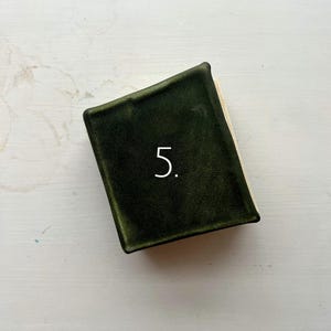 May include: A small, dark green book with a textured cover. The number "5" is printed in white on the cover. The book's pages are visible on the right side.