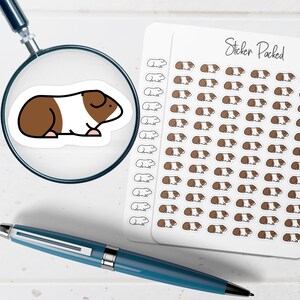 May include: A sheet of small stickers featuring brown and white guinea pig illustrations, with the text Sticker Packed at the top. A magnifying glass shows a larger view of one guinea pig sticker, and a blue pen lies horizontally below the sheet.