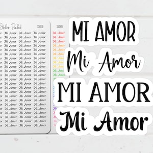 May include: A collection of white stickers featuring the phrase "Mi Amor" in black, cursive lettering. Several stickers are visible, with the phrase repeated in a smaller font. The stickers are set against a light backdrop.