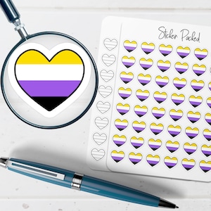 May include: A heart-shaped sticker with the non-binary pride flag colors of yellow, white, purple, and black, viewed through a magnifying glass. A sheet of heart stickers with the same design is visible. A blue pen rests on the white surface.