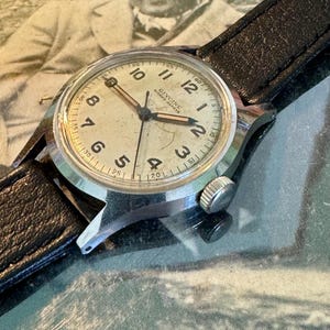 May include: Vintage Glycine watch with a silver-tone case and a black leather band. The watch face is off-white with black numerals and hands. The watch has a small second hand and the brand name is visible. The background is a vintage photograph.