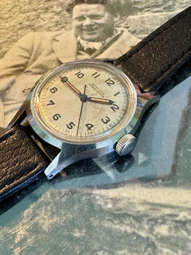 May include: Vintage Glycine watch with a silver-tone case and a black leather band. The watch face is off-white with black numerals and hands. The watch has a small second hand and the brand name is visible. The background is a vintage photograph.