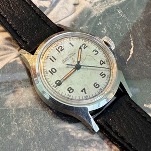 May include: Vintage Glycine watch with a silver-tone case and a black leather band. The watch face is off-white with black numerals and hands. The watch has a second hand and the brand name "Glycine" is visible on the face.