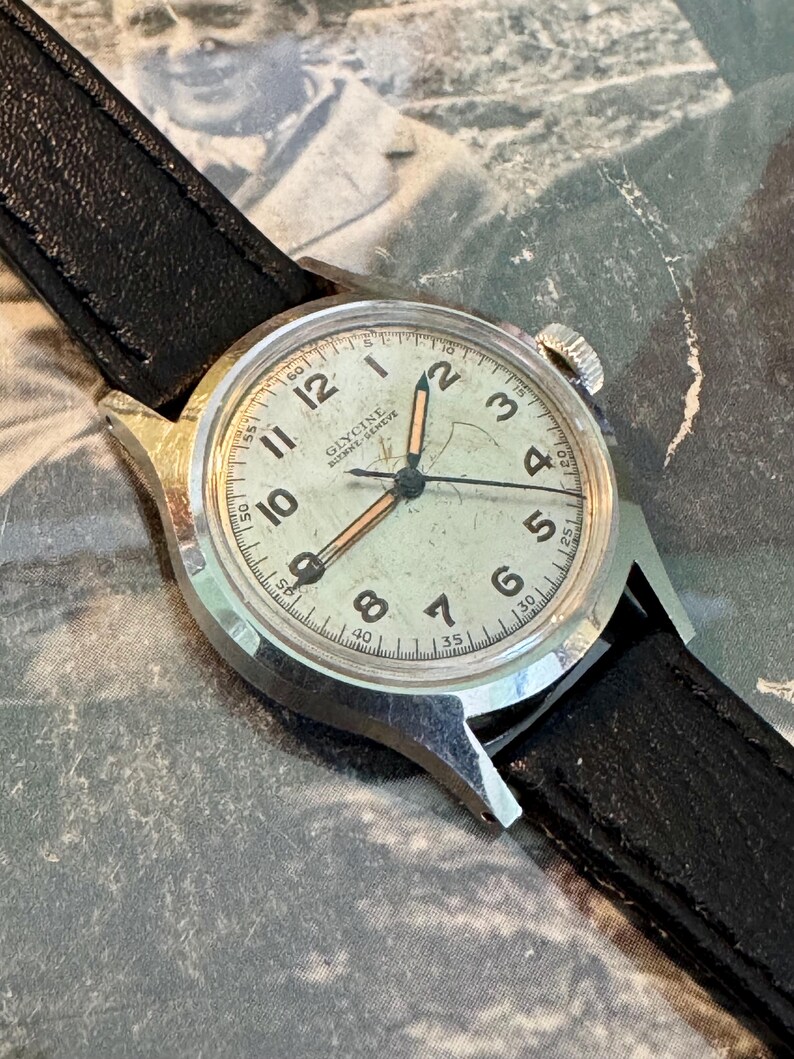 May include: Vintage Glycine watch with a silver-tone case and a black leather band. The watch face is off-white with black numerals and hands. The watch has a second hand and the brand name "Glycine" is visible on the face.