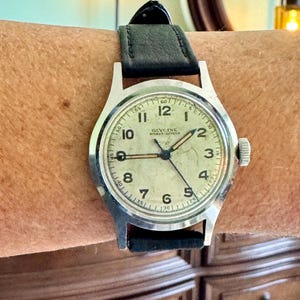 May include: A vintage Glycine wristwatch with a silver-colored case and a black leather band. The watch face is off-white with black numerals and hands. The watch is on a wrist.