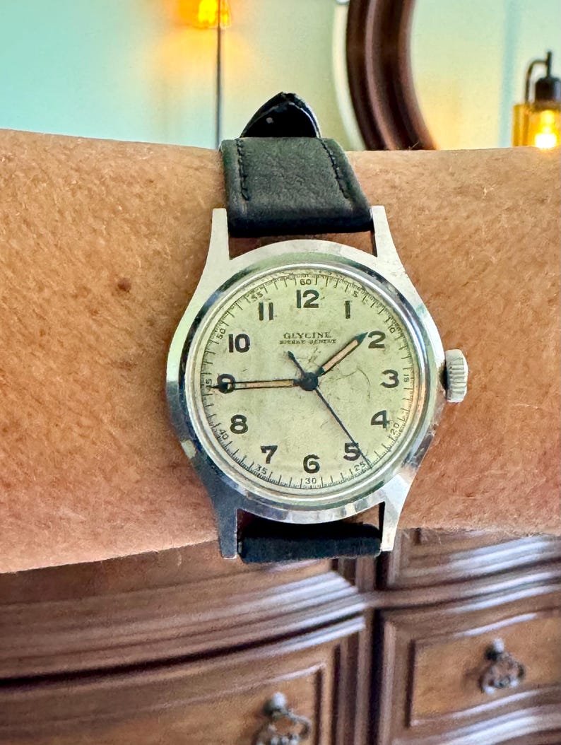 May include: A vintage Glycine wristwatch with a silver-colored case and a black leather band. The watch face is off-white with black numerals and hands. The watch is on a wrist.