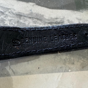 May include: Close-up of a black leather strap with the embossed text "GENUINE LEATHER". The strap appears to be from a watch or similar accessory. The background is a faded, textured surface.