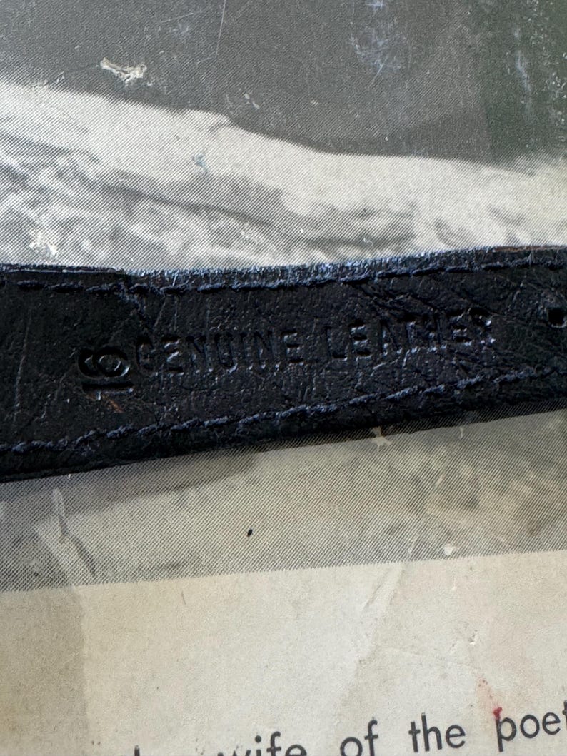 May include: Close-up of a black leather strap with the embossed text "GENUINE LEATHER". The strap appears to be from a watch or similar accessory. The background is a faded, textured surface.