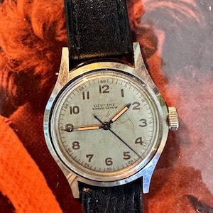 May include: A vintage Glycine Bienne-Geneve wristwatch with a silver-tone case and a black leather strap. The watch face is light green with black numerals and hands. The watch has a crown on the right side.