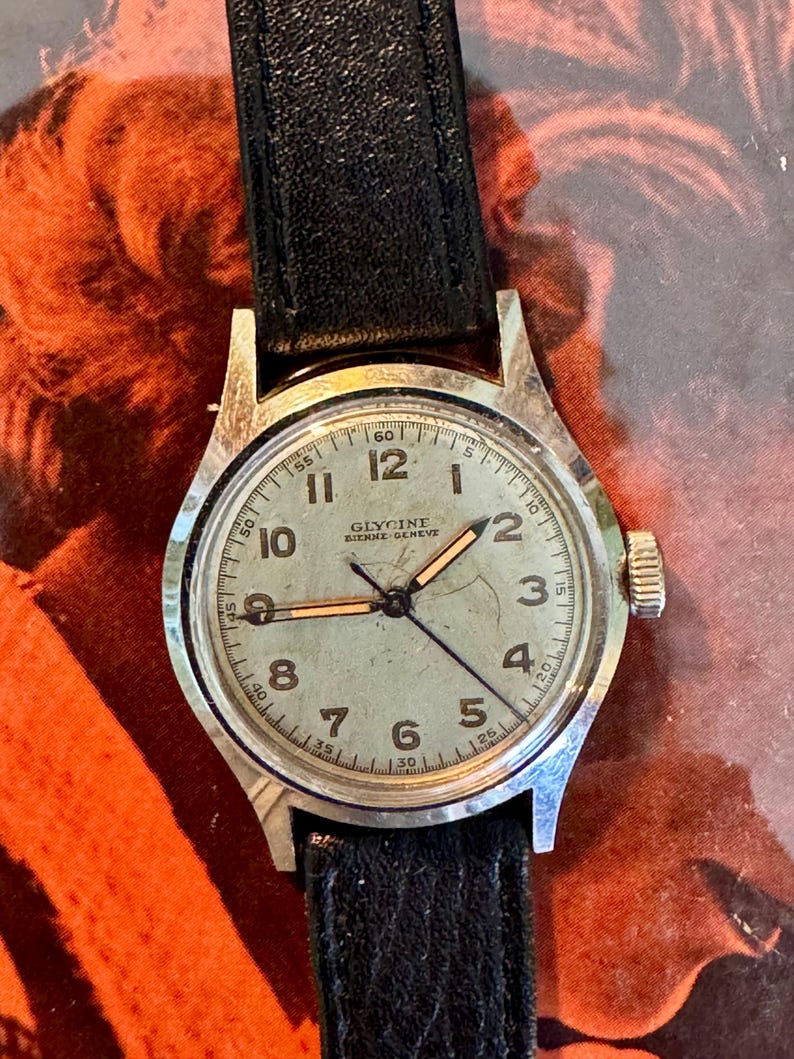 May include: A vintage Glycine Bienne-Geneve wristwatch with a silver-tone case and a black leather strap. The watch face is light green with black numerals and hands. The watch has a crown on the right side.