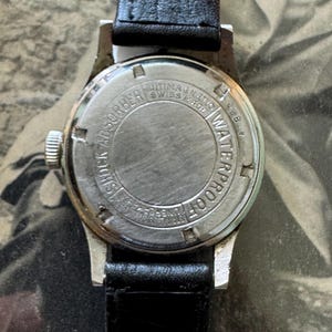May include: A close-up of the back of a vintage wristwatch with a black leather band. The watch case is silver-toned with the words "SHOCK ABSORBER", "ANTIMAGNETIC", "SWISS MADE", and "WATERPROOF" engraved on the back.