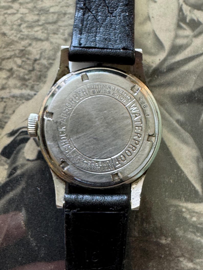 May include: A close-up of the back of a vintage wristwatch with a black leather band. The watch case is silver-toned with the words "SHOCK ABSORBER", "ANTIMAGNETIC", "SWISS MADE", and "WATERPROOF" engraved on the back.
