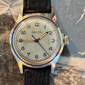 May include: Vintage Glycine watch with a silver-tone case and a black leather band. The watch face is light green with black numerals and hands. The watch has a small crown and the brand name "Glycine" is visible.