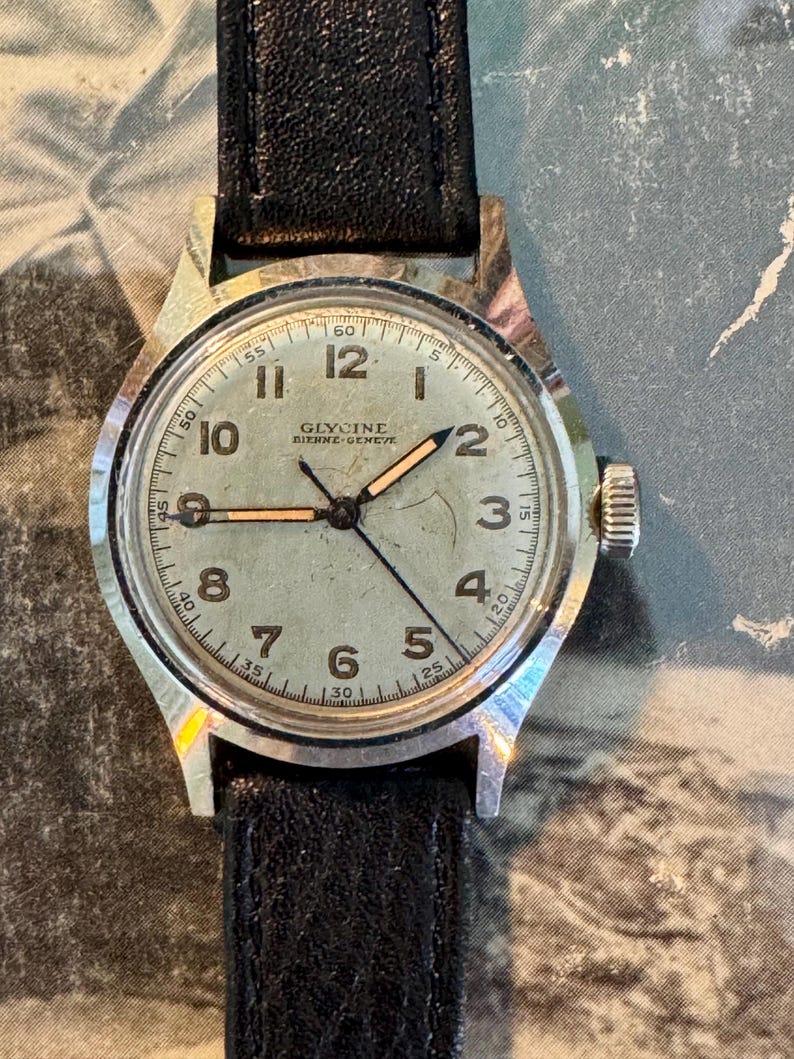 May include: Vintage Glycine watch with a silver-tone case and a black leather band. The watch face is light green with black numerals and hands. The watch has a small crown and the brand name "Glycine" is visible.