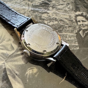 May include: Close-up of a vintage wristwatch with a black leather band. The watch face is silver with black hands and markers. The back of the watch is stainless steel and reads "Waterproof" and "Shock Absorber".