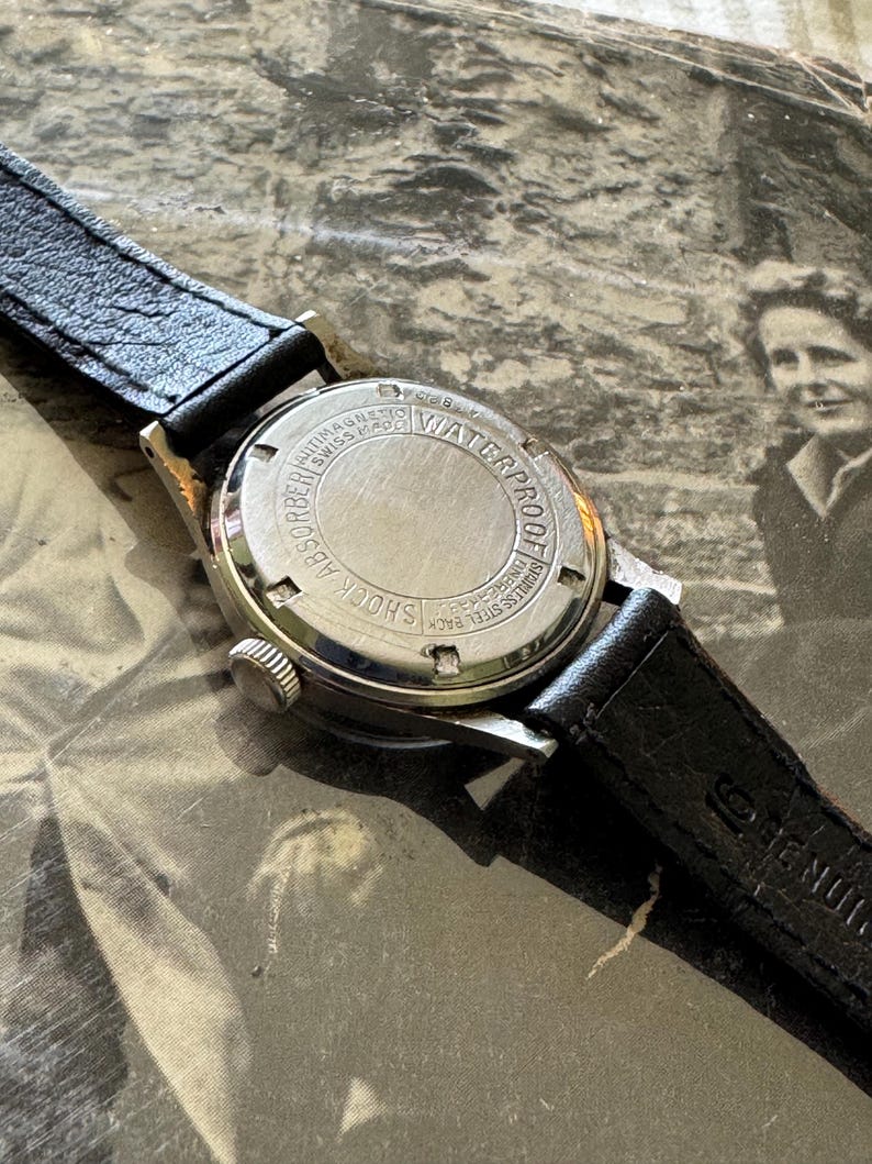 May include: Close-up of a vintage wristwatch with a black leather band. The watch face is silver with black hands and markers. The back of the watch is stainless steel and reads "Waterproof" and "Shock Absorber".