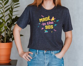 May include: A navy blue t-shirt with a graphic print that says "MADE in the 90s" in yellow, pink, and blue. The graphic is surrounded by colorful geometric shapes.