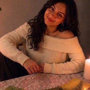 May include: An off-the-shoulder cream-colored ribbed knit sweater with long sleeves. The garment features a wide folded collar that rests across the shoulders and upper chest, providing a textured appearance with vertical ribbing throughout the fabric.