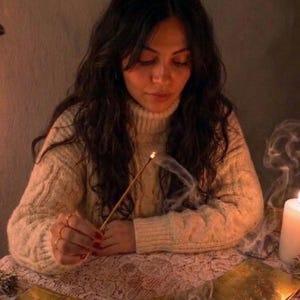 May include: A person wearing a cream-colored cable knit turtleneck sweater holds a lit incense stick with visible smoke. A row of gold-toned rectangular cards is arranged on a white lace tablecloth next to a white pillar candle.