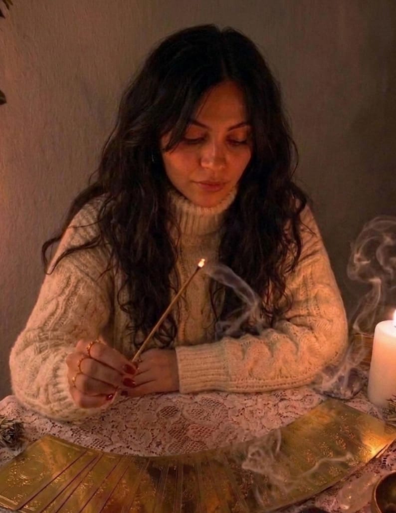 May include: A person wearing a cream-colored cable knit turtleneck sweater holds a lit incense stick with visible smoke. A row of gold-toned rectangular cards is arranged on a white lace tablecloth next to a white pillar candle.
