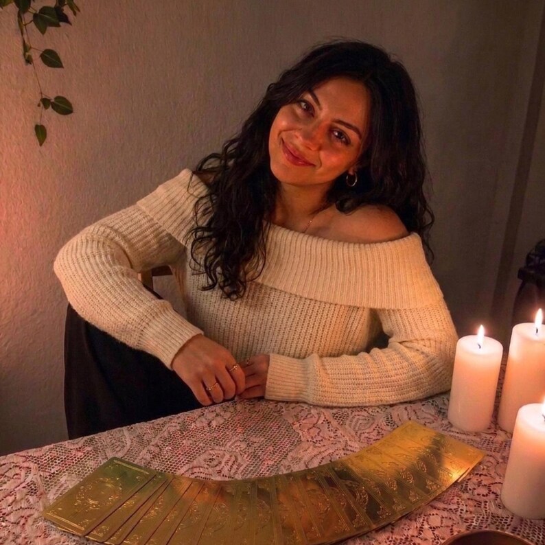 A woman in a white sweater sits at a table with candles and tarot cards, smiling at the camera.