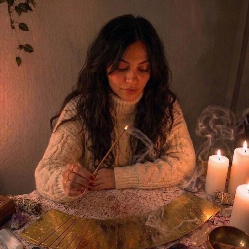 A woman sits at a table, holding a stick and surrounded by candles, tarot cards, and crystals, appearing to be engaged in a ritual or ceremony.
