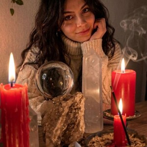 A woman with long dark hair is sitting at a table with candles, crystals, and a crystal ball, surrounded by a potted plant and a sign that reads "House of Magic Miracles".