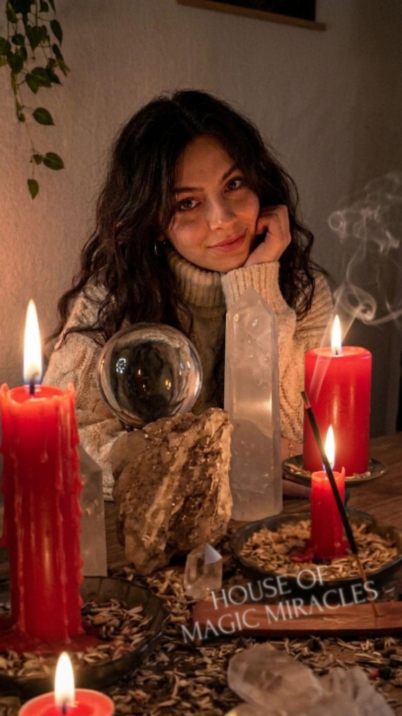 A woman with long dark hair is sitting at a table with candles, crystals, and a crystal ball, surrounded by a potted plant and a sign that reads "House of Magic Miracles".